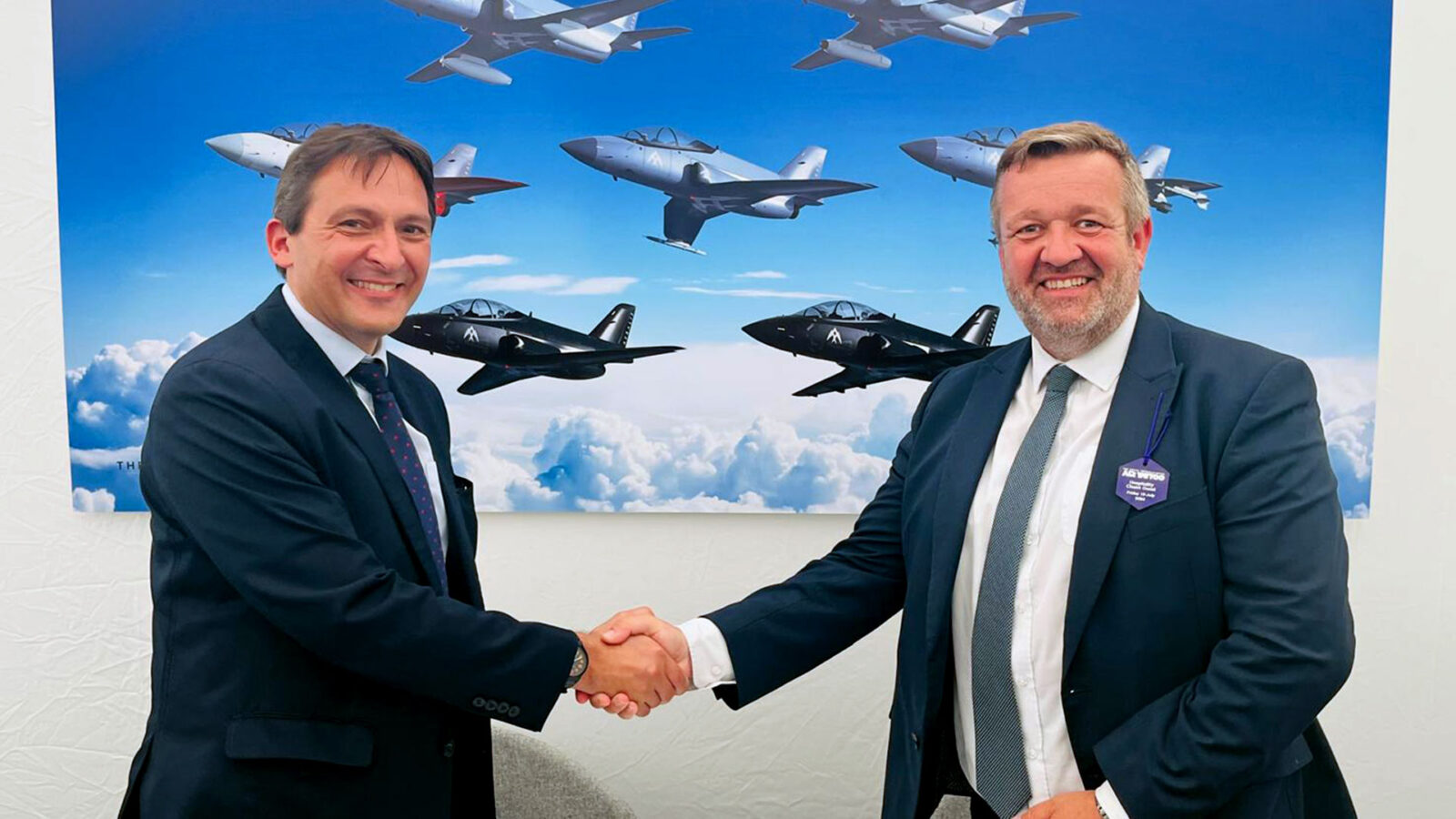 Tristan Crawford, CEO and Founder of AERALIS is pictured shaking hands with Andrew Ferguson, VP Sales and Business Development, EMEA and APAC for StandardAero, in front of a large picture of a formation of seven AERALIS jets in different mission configurations.