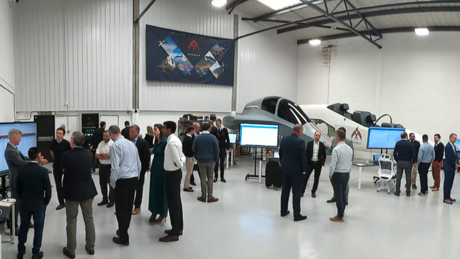 A group of people in a large indoor space with screens and a helicopter cockpit simulator, surrounded by banners and white walls.